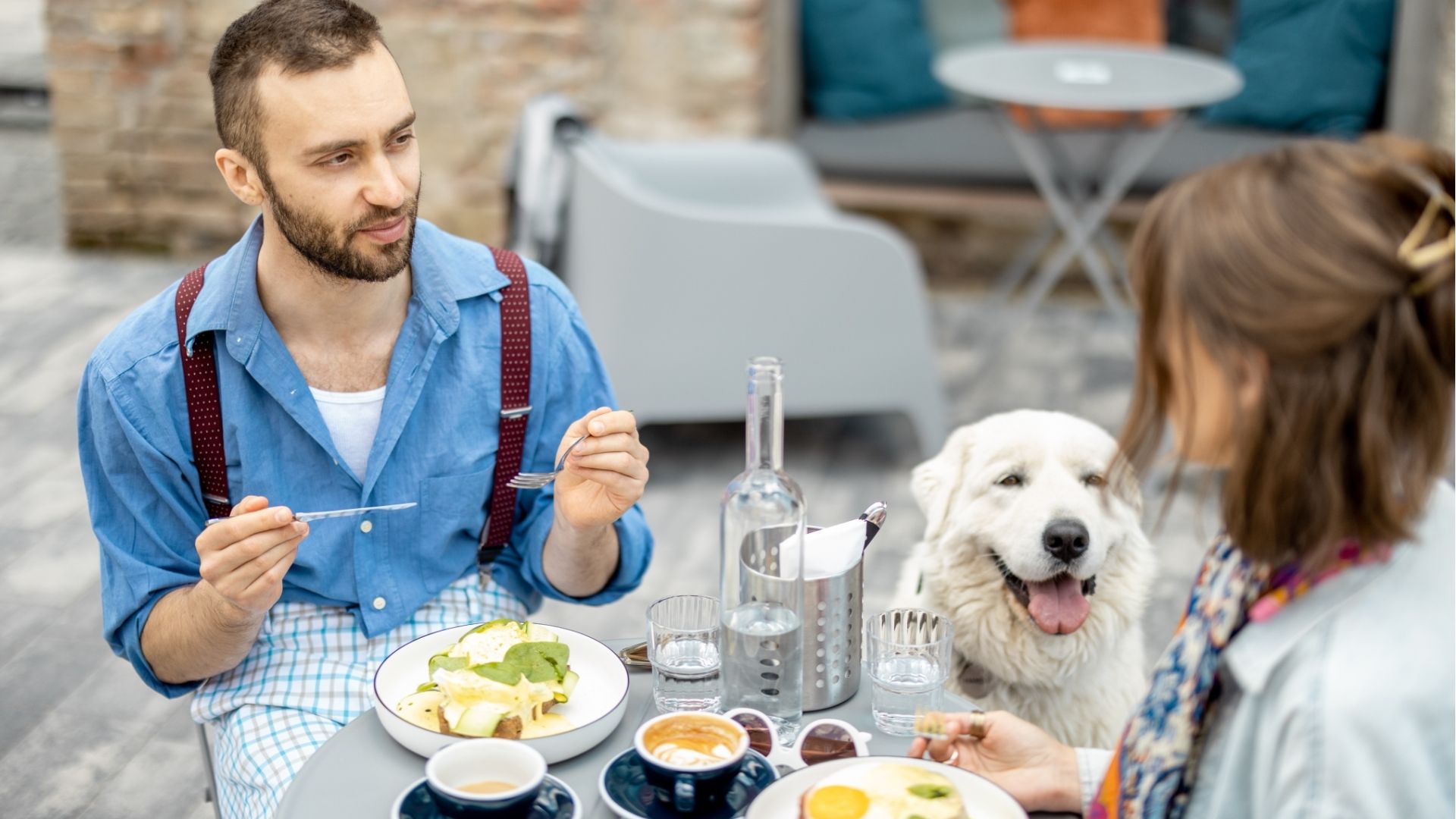Stylish man and woman have a breakfast while sitting with dog at cafe outdoors