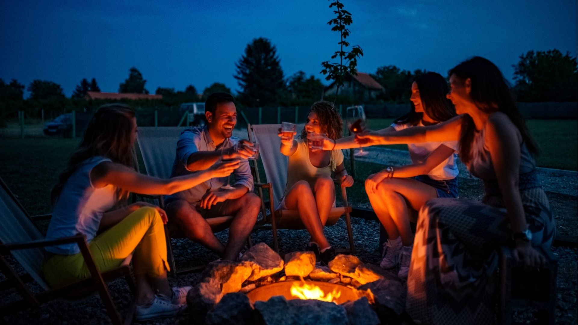 Friends enjoying evening drinks by firepit outdoors.
