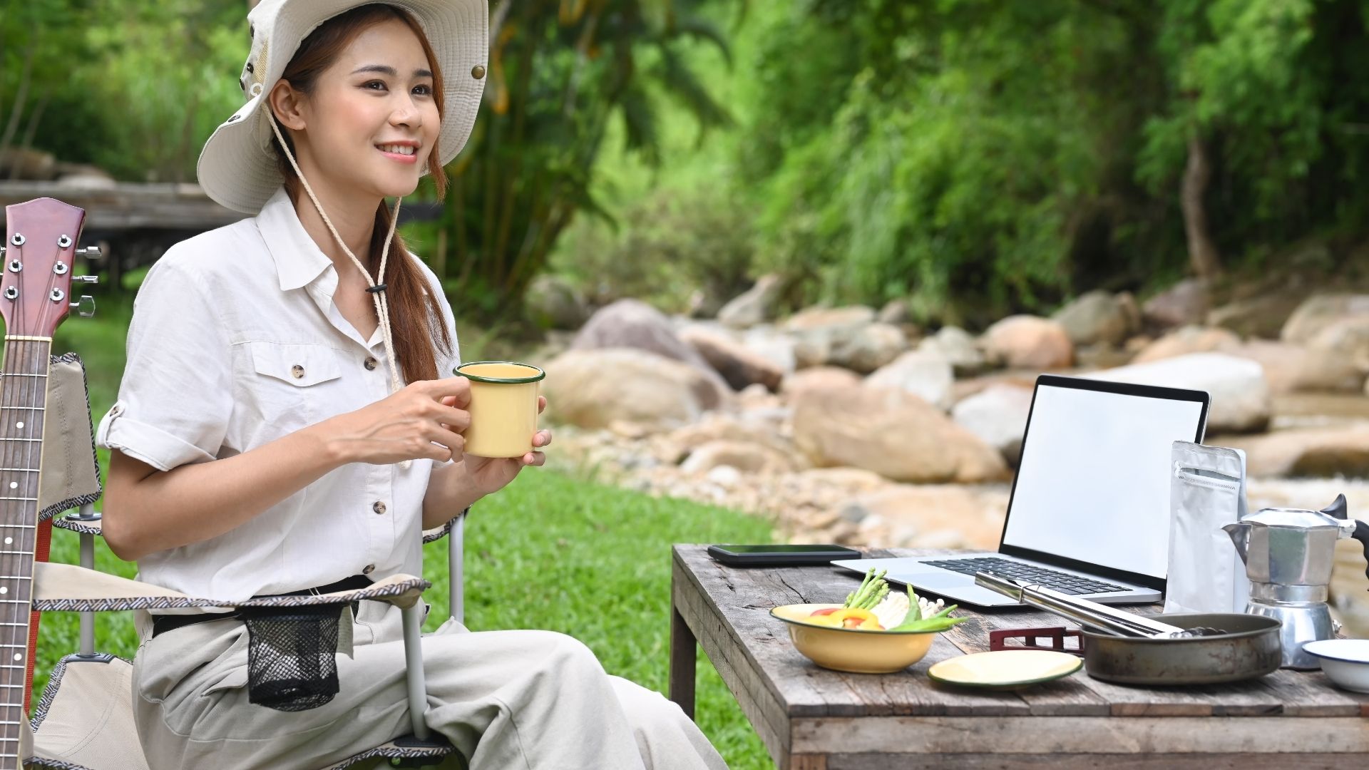 Traveler sitting with a cup near a laptop and camping setup by a stream.