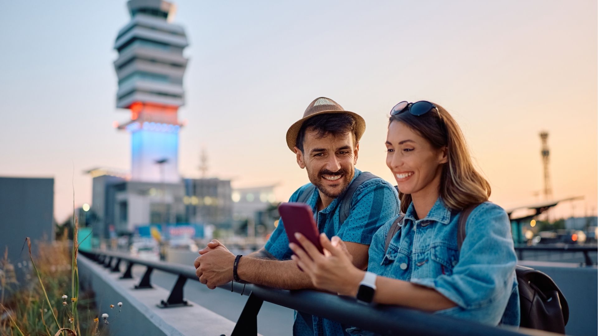 Happy couple of tourists using cell phone at the airport at sunset.