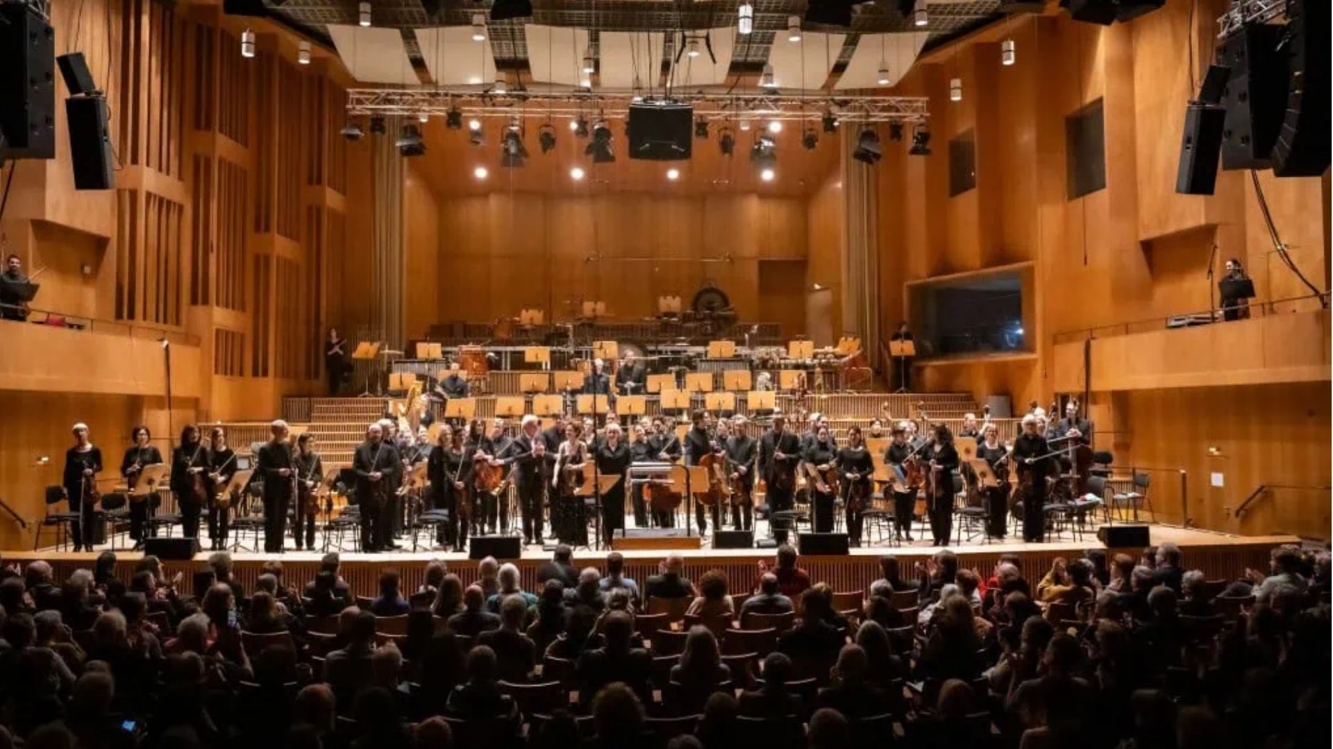 Orchestra taking a bow onstage in a wooden concert hall before a seated audience.