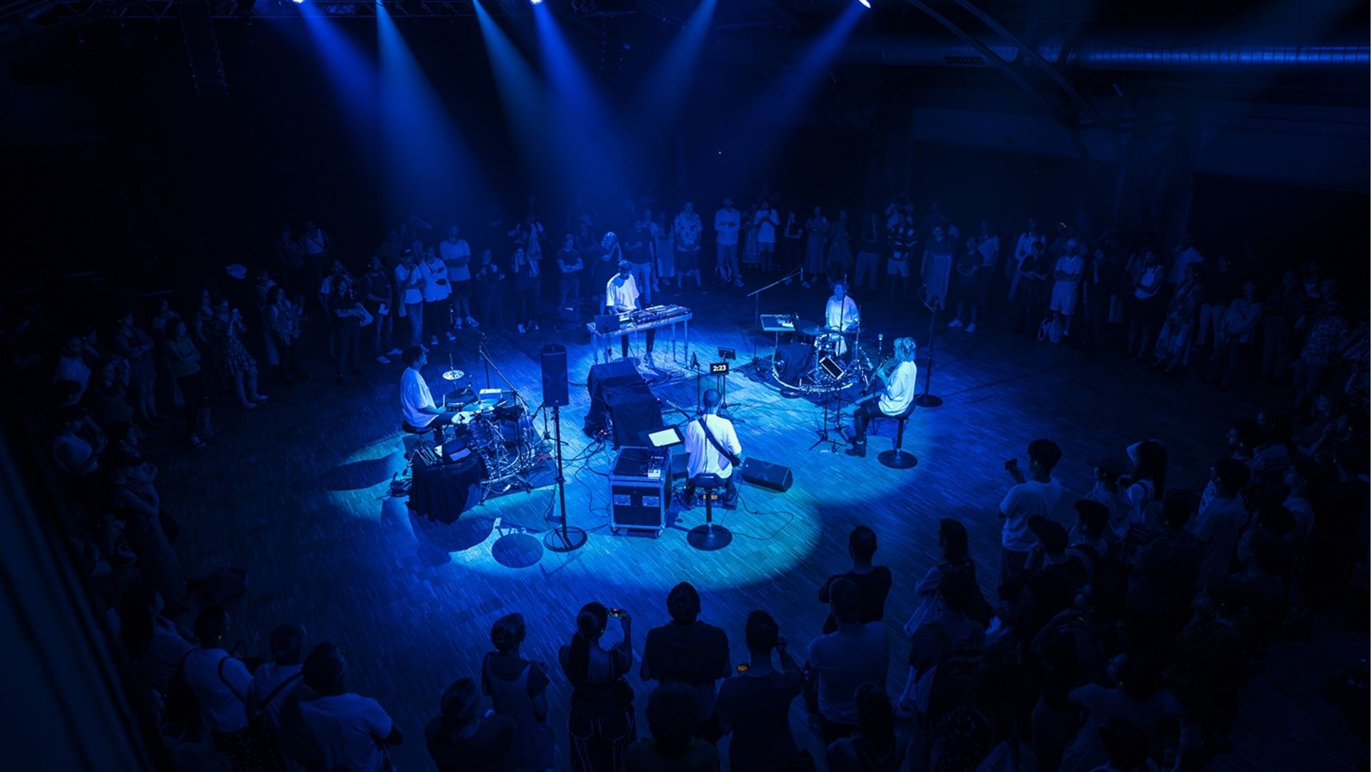 Musicians perform in a blue-lit circular stage surrounded by a standing audience