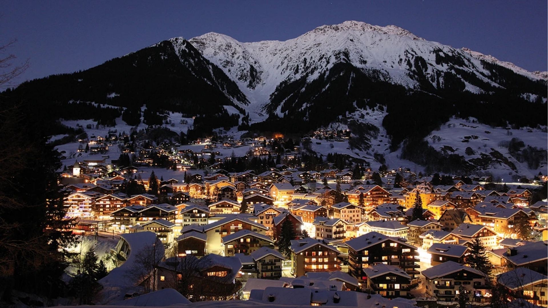 Snow covered alpine town illuminated at night with mountains in the background during winter