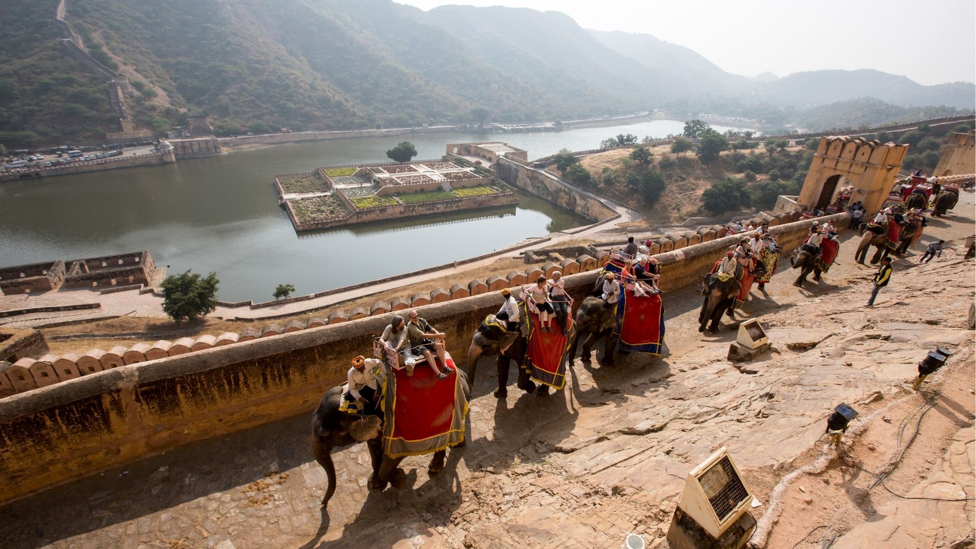 Tourists riding elephants on a scenic path overlooking a historic fortress and water reservoir